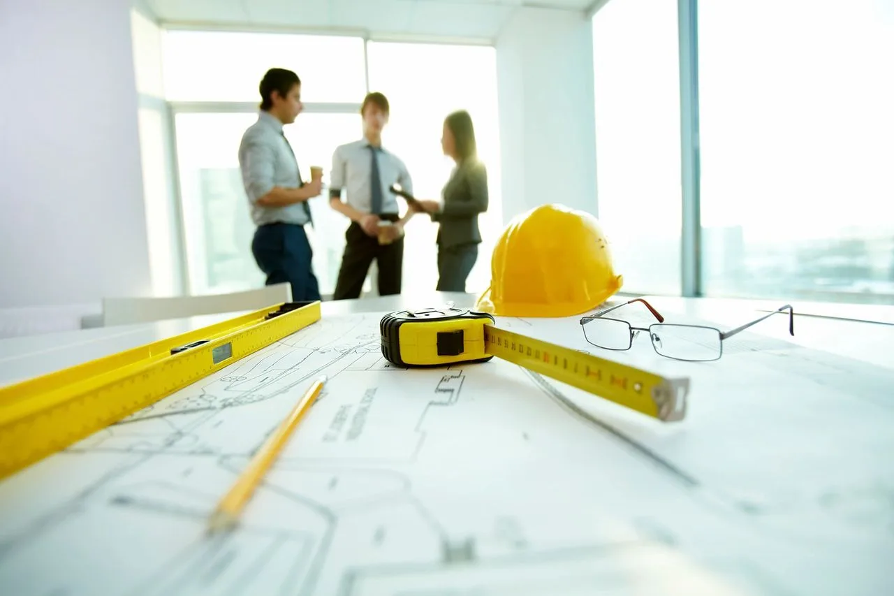 Blueprints, a hard hat, glasses, and a tape measure laid out on a table with colleagues talking in the background