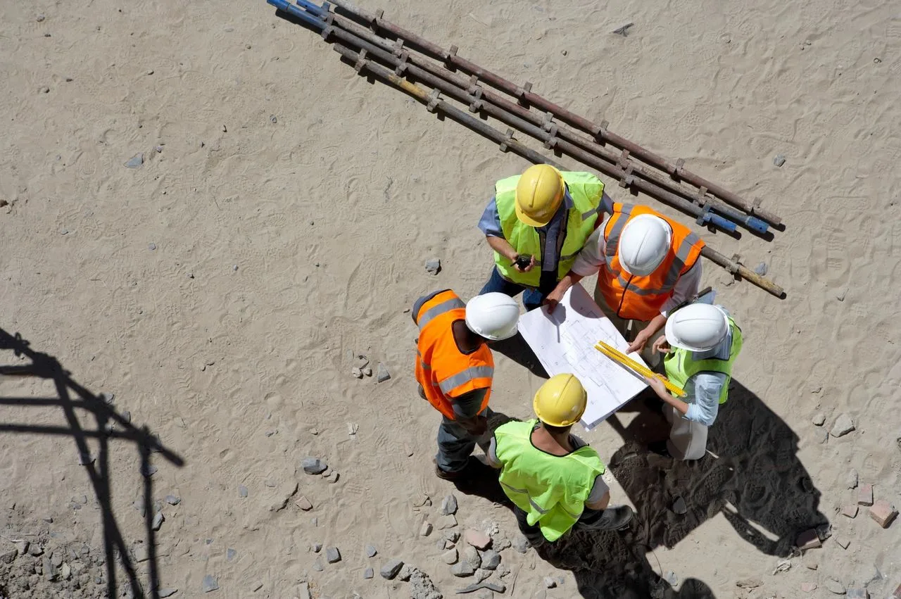 Five construction workers in hard hats gather around blueprints on a dirt job site