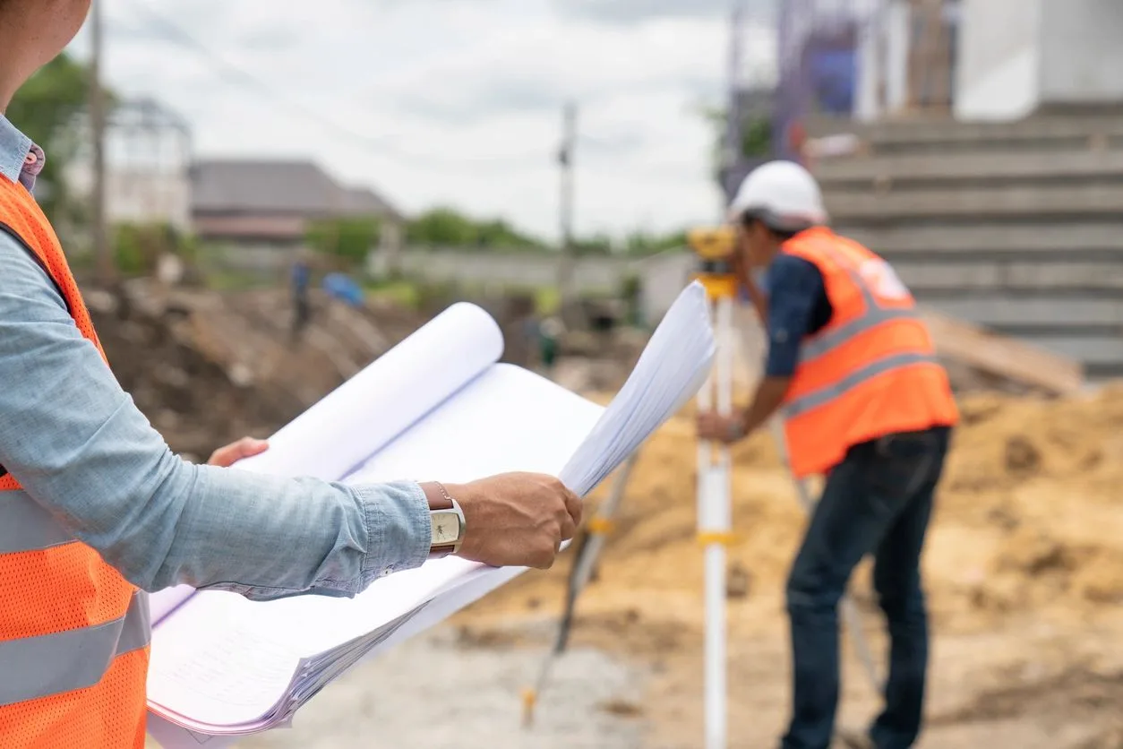 Construction consultant holding blueprints while a surveyor works in the background