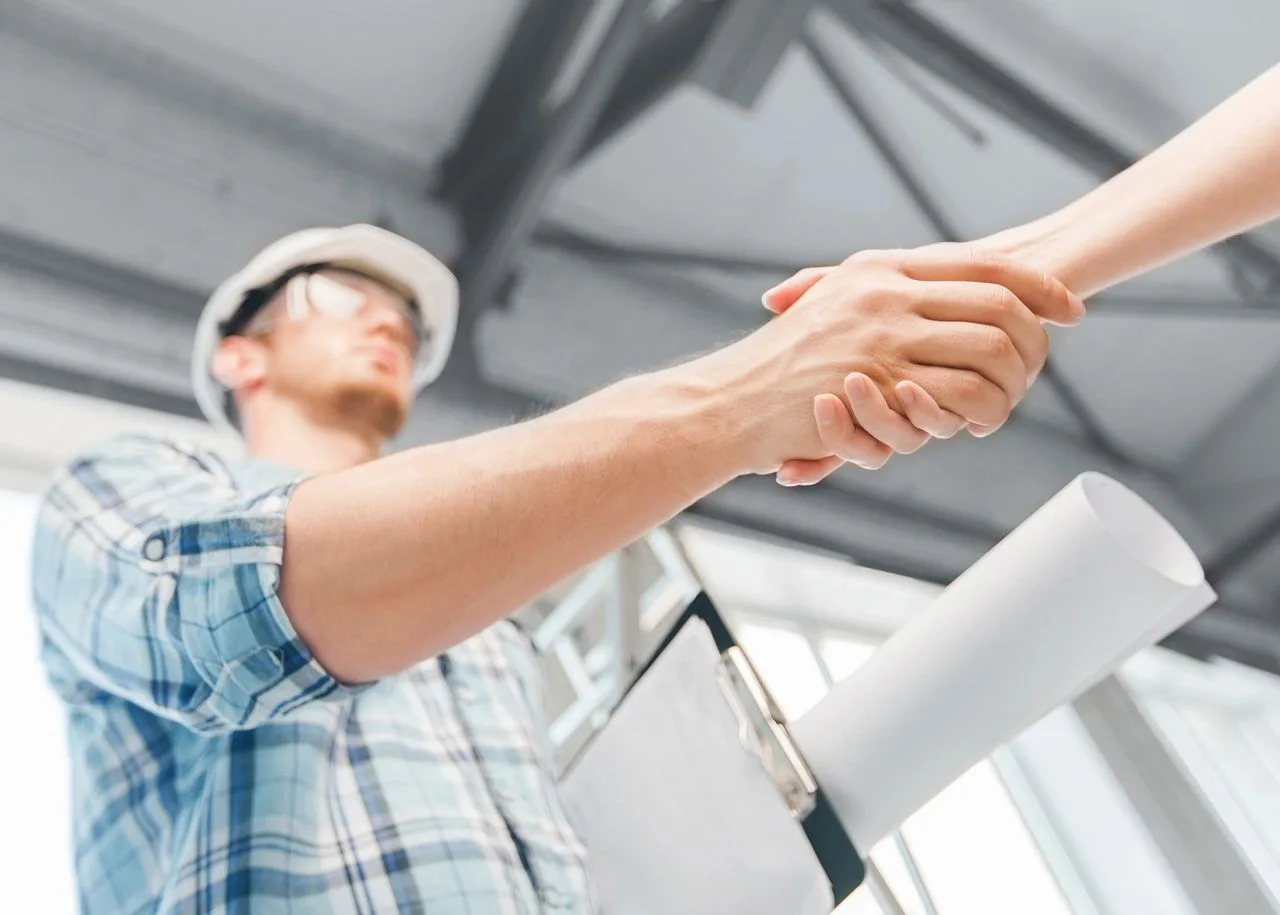 Contractor in a hard hat shaking hands with a client while holding blueprints