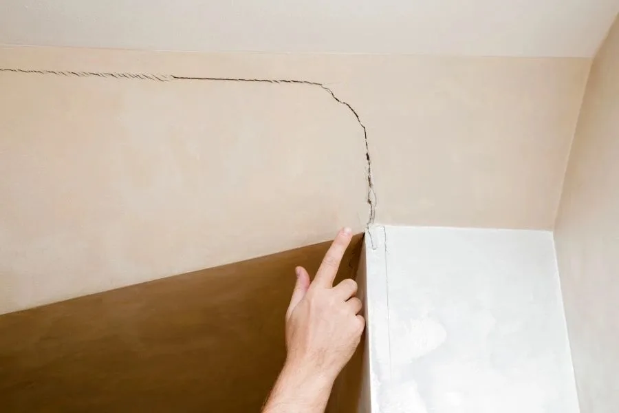 Hand pointing to a diagonal crack running along a ceiling corner, a sign of a construction defect