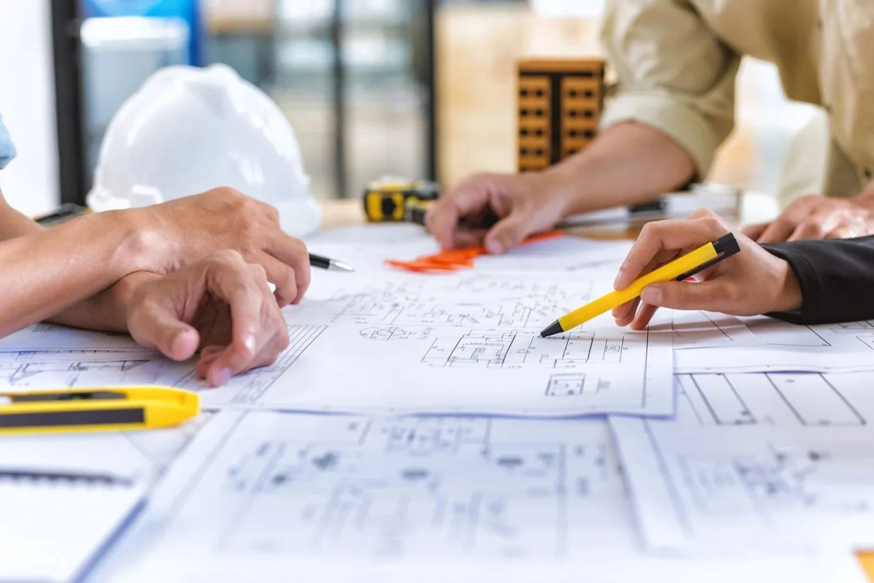 Hands reviewing building blueprints with a hard hat and tape measure on the table
