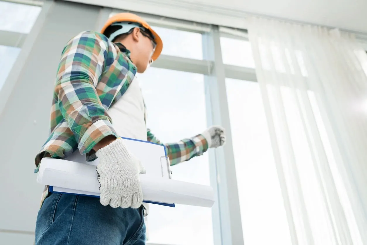 Inspector in a hard hat holding a clipboard while examining large windows in a home