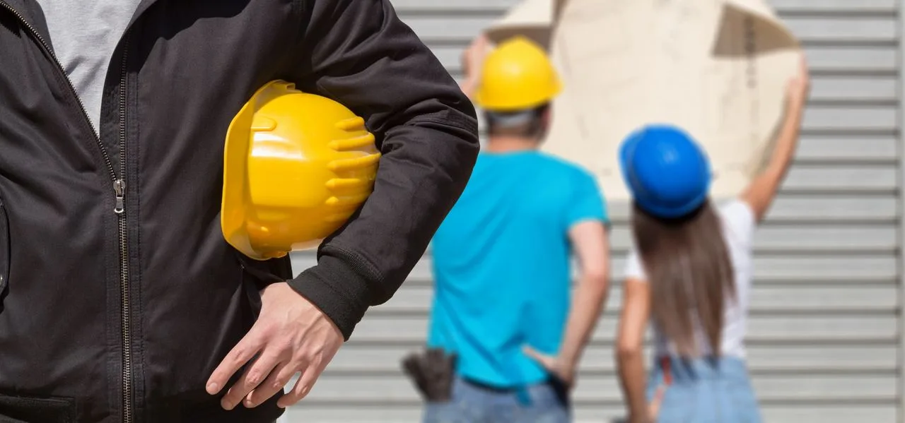 Man holding a yellow hard hat while colleagues review a blueprint in the background