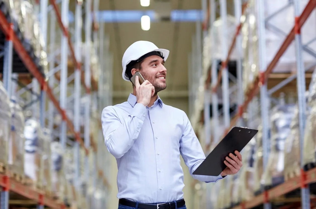 Man in a hard hat talking on the phone while holding a clipboard in a warehouse
