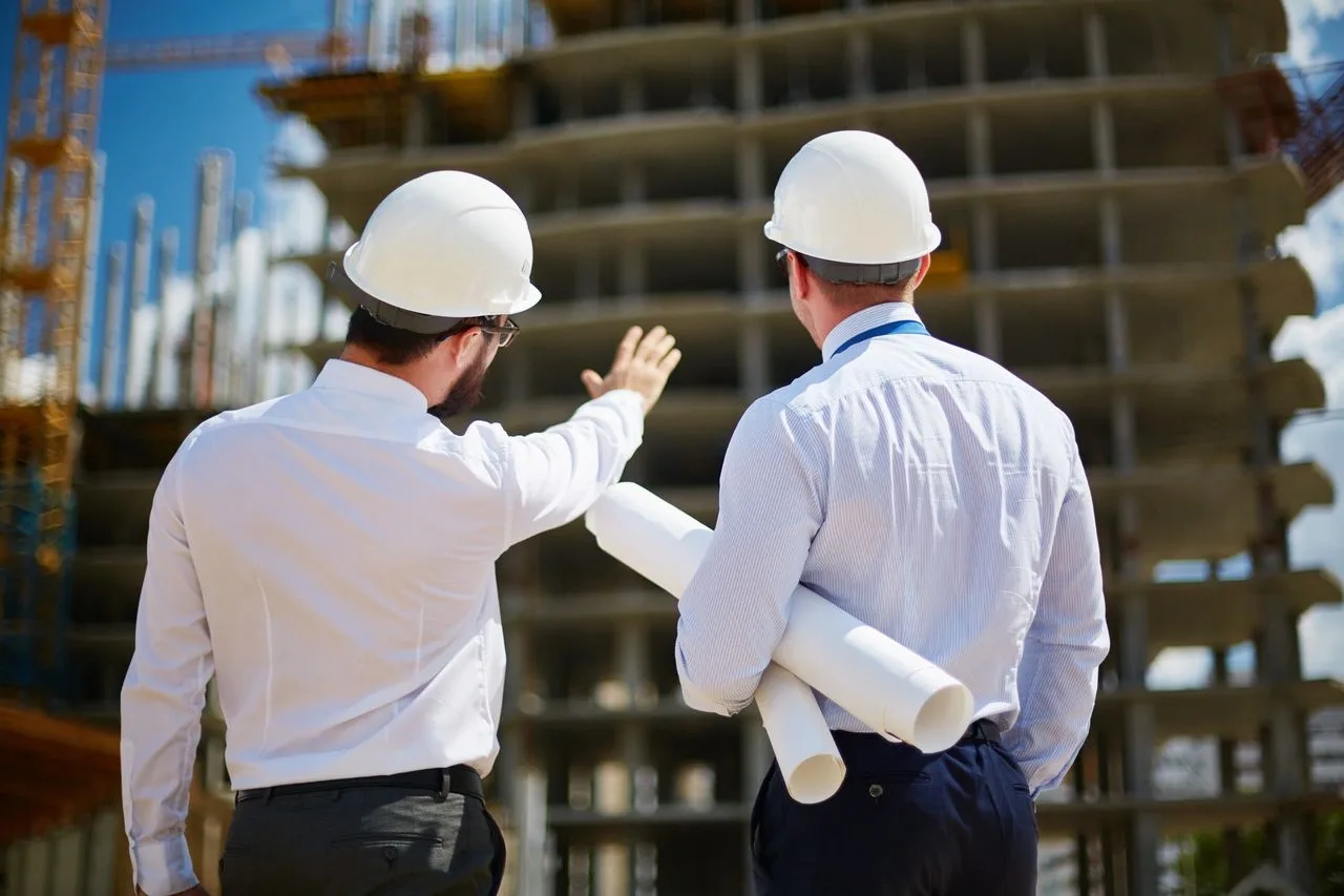 Two men in hard hats reviewing a high-rise construction site, one pointing while the other holds rolled plans