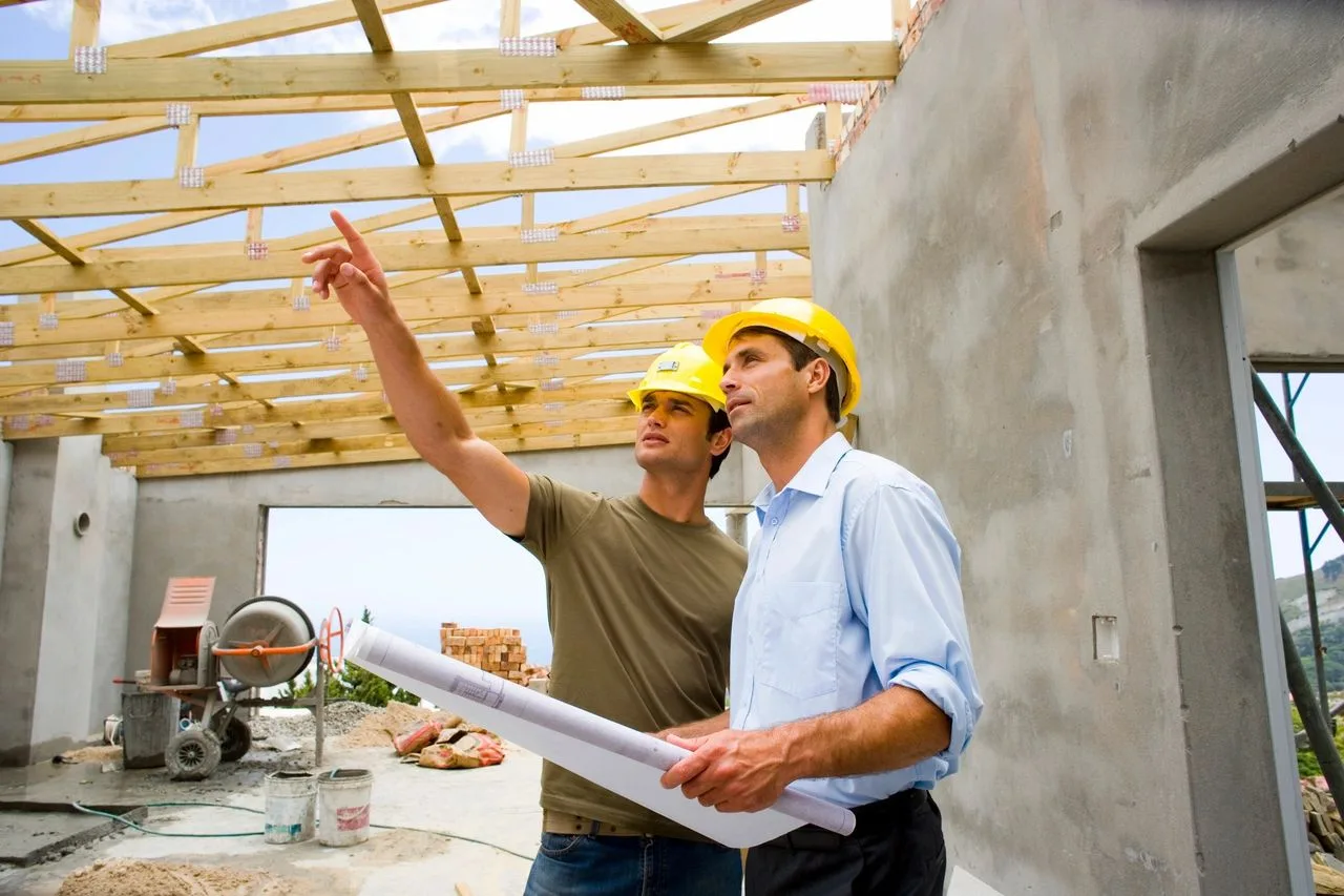 Two men reviewing plans beneath open roof trusses at a home under construction