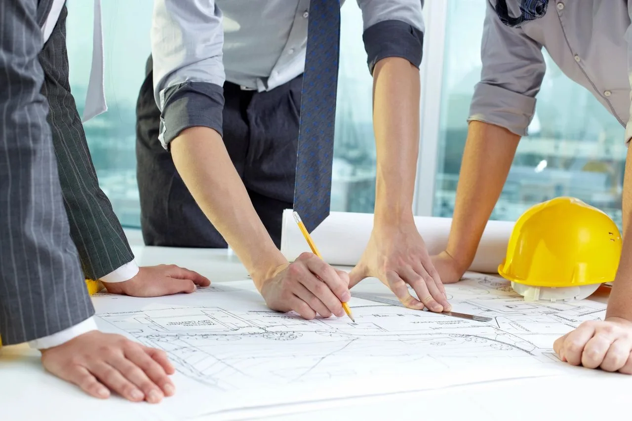 Team reviewing blueprints on a table, one person pointing with a pencil, a hard hat beside the plans