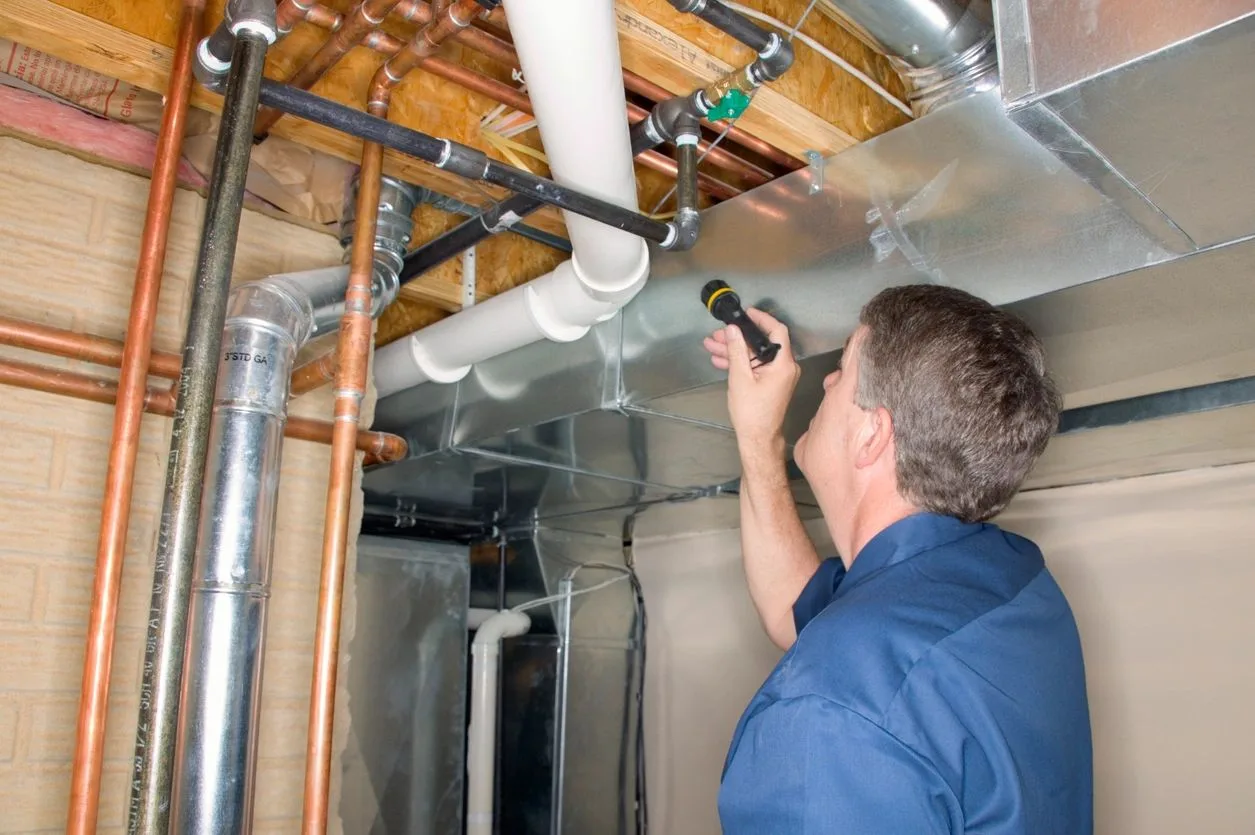 Technician using a flashlight to inspect ductwork and pipes above a ceiling