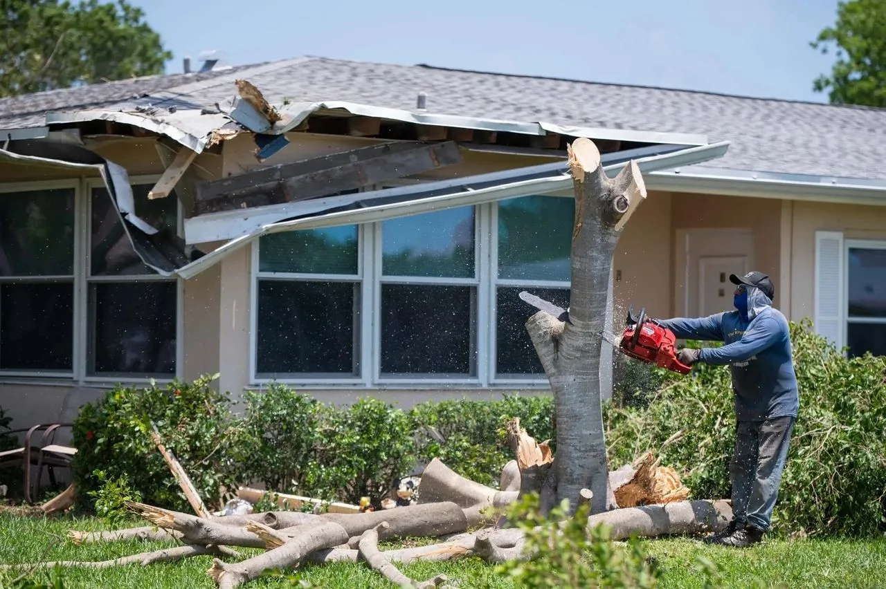 Man uses a chainsaw to clear a fallen tree beside a home with storm-damaged roof after the Palm Beach Gardens tornado