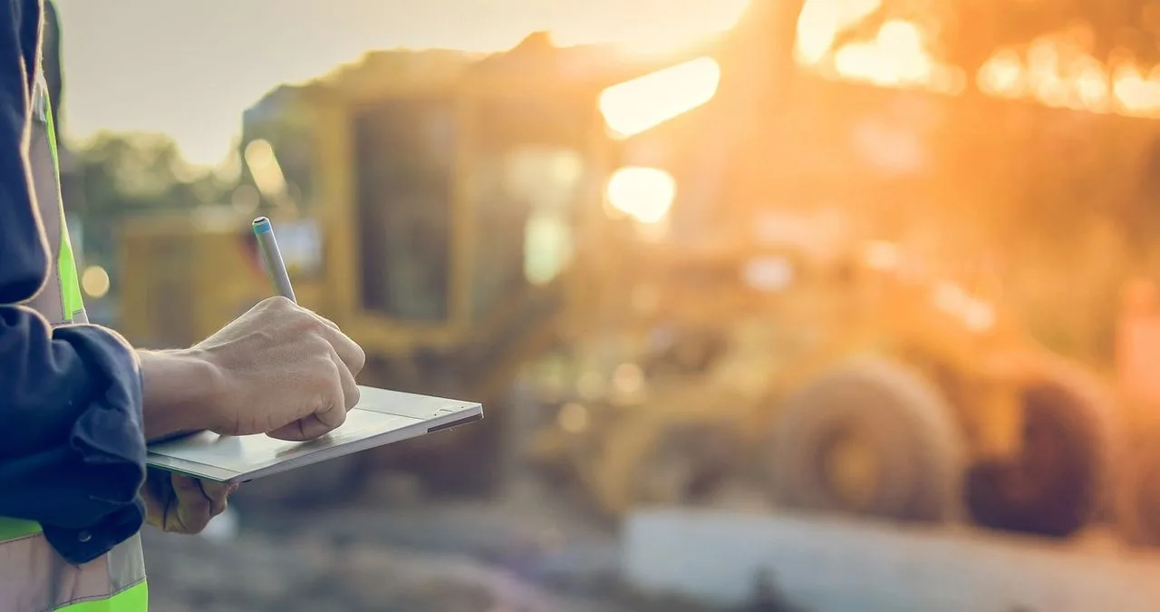 Worker in a safety vest taking notes on a clipboard at a job site with heavy equipment