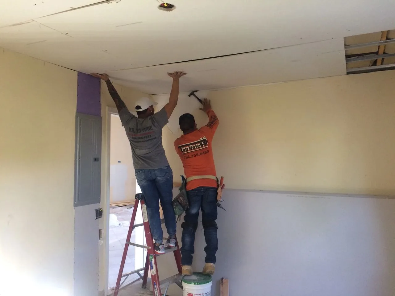 Two workers installing drywall on a ceiling with a ladder and tools