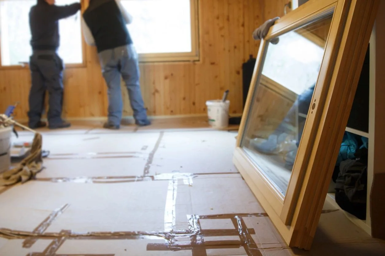 Two workers installing a new window in a wood-paneled room with the floor covered in tarps