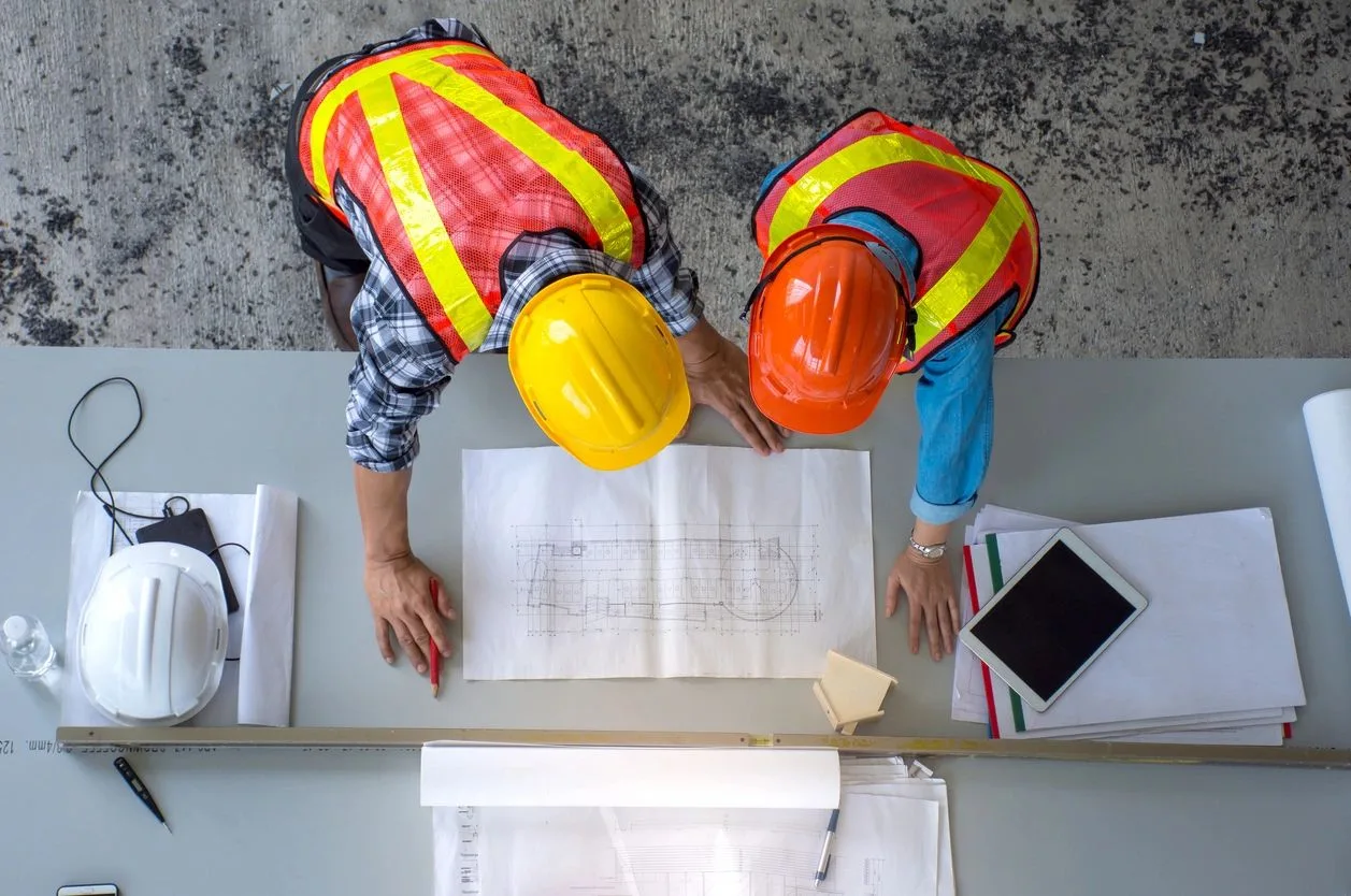 Two workers in hard hats reviewing blueprints on a table, seen from above